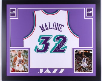 May include: A framed white basketball jersey with the name "MALONE" and the number "32" in purple and teal. The jersey is signed by Karl Malone. The frame has two smaller photos of Malone playing basketball.