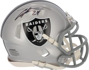 May include: Silver football helmet with the Raiders logo, a black and white emblem featuring a pirate. The helmet has a gray face mask and a signature with the number 24. The helmet is a collectible item.