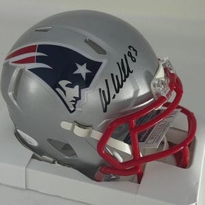 Puede incluir: Casco de f&uacute;tbol americano plateado con una m&aacute;scara roja. El casco presenta el logotipo del equipo en rojo, blanco y azul, y una firma con tinta negra. El casco est&aacute; en una caja blanca.