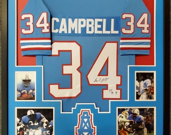 Puede incluir: Una camiseta de fútbol azul enmarcada con el número 34 y el nombre "Campbell" en la espalda. La camiseta está firmada por el jugador e incluye fotos del jugador en acción.