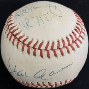 Pode incluir: Uma bola de beisebol branca assinada pelo membro do Hall da Fama, Hank Aaron. A assinatura está em tinta azul e diz "Hank Aaron". A bola de beisebol também tem uma inscrição em tinta azul que diz "Apr. 7, 1975".