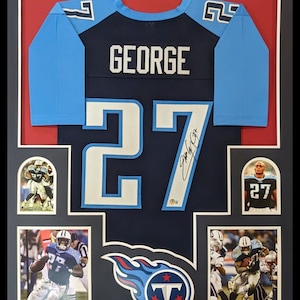 May include: A framed autographed football jersey with the name "GEORGE" and the number "27" on the back. The jersey is blue with light blue accents and the Tennessee Titans logo. The frame includes three smaller photos of the player in action.