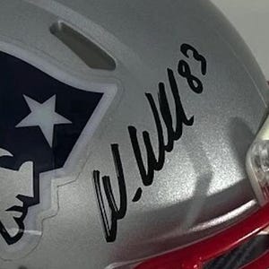 Puede incluir: Casco de f&uacute;tbol americano plateado con el logotipo de los New England Patriots y una firma. El logotipo presenta una estrella blanca y la silueta de una cabeza de patriota sobre un fondo azul marino. El casco tiene una m&aacute;scara roja.