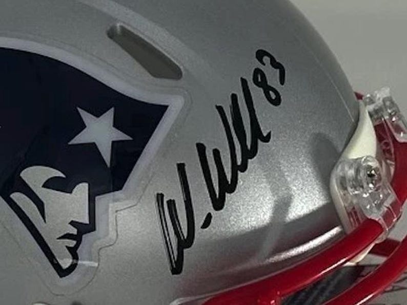 Puede incluir: Casco de f&uacute;tbol americano plateado con el logotipo de los New England Patriots y una firma. El logotipo presenta una estrella blanca y la silueta de una cabeza de patriota sobre un fondo azul marino. El casco tiene una m&aacute;scara roja.
