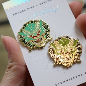 Puede incluir: Dos pines de esmalte en tono dorado con un diseño estilizado de cabeza de león en verde y dorado. Los pines están en una tarjeta blanca con el texto "EYESWHISKERS.COM".
