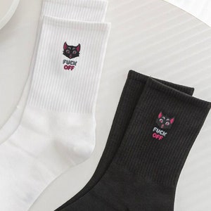 May include: A pair of socks, one white and one black, with a black cat graphic and the text "F*CK OFF" in pink lettering.