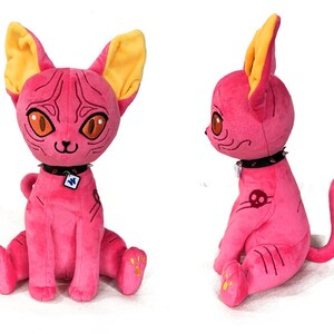 May include: Pink plush cat toy with yellow ears, a black studded collar, and a skull tattoo. The cat is sitting with its tail curled behind it.