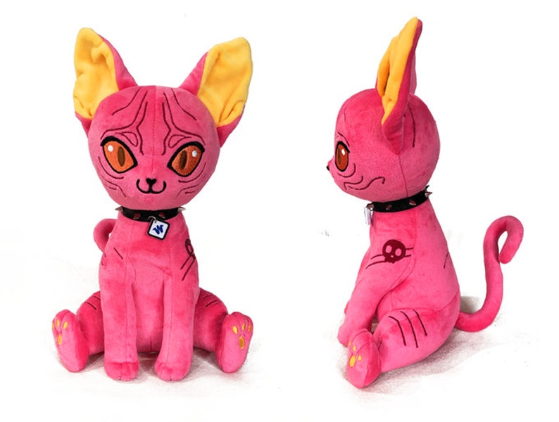 May include: Pink plush cat toy with yellow ears, a black studded collar, and a skull tattoo. The cat is sitting with its tail curled behind it.