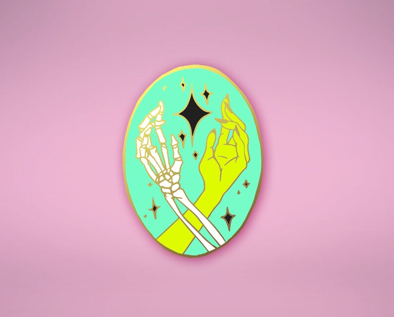May include: A gold-colored enamel pin with a turquoise background. The pin features a skeleton hand and a flesh hand reaching towards each other, with a black star in the center. The background is dotted with small gold stars.