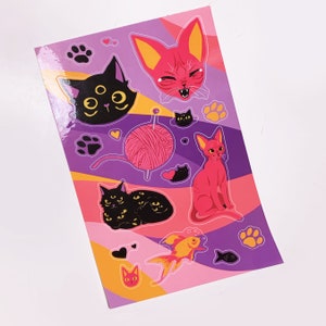 May include: A sheet of stickers featuring various cats and kittens. The stickers are on a purple background with pink and yellow accents. The cats are black, pink, and orange. There are also stickers of a ball of yarn, a fish, and paw prints.