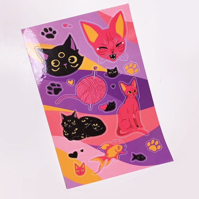May include: A sheet of stickers featuring various cats and kittens. The stickers are on a purple background with pink and yellow accents. The cats are black, pink, and orange. There are also stickers of a ball of yarn, a fish, and paw prints.