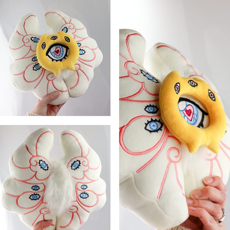 Puede incluir: Almohada de felpa blanca con bordado rosa y rojo que representa un ojo estilizado con un coraz&oacute;n en el centro. La almohada tiene un anillo de felpa amarillo con tres ojos y un coraz&oacute;n en el centro.