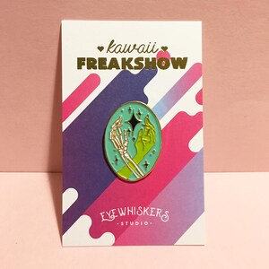 May include: A teal enamel pin with a gold outline featuring a skeleton hand and a green hand holding a star. The pin is on a white card with the text "kawaii FREAKSHOW" in gold lettering and "EYE WHISKERS STUDIO" in black lettering.