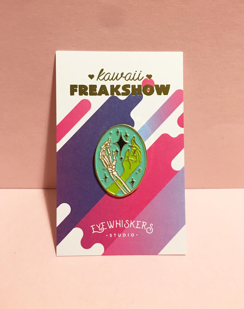 May include: A teal enamel pin with a gold outline featuring a skeleton hand and a green hand holding a star. The pin is on a white card with the text "kawaii FREAKSHOW" in gold lettering and "EYE WHISKERS STUDIO" in black lettering.