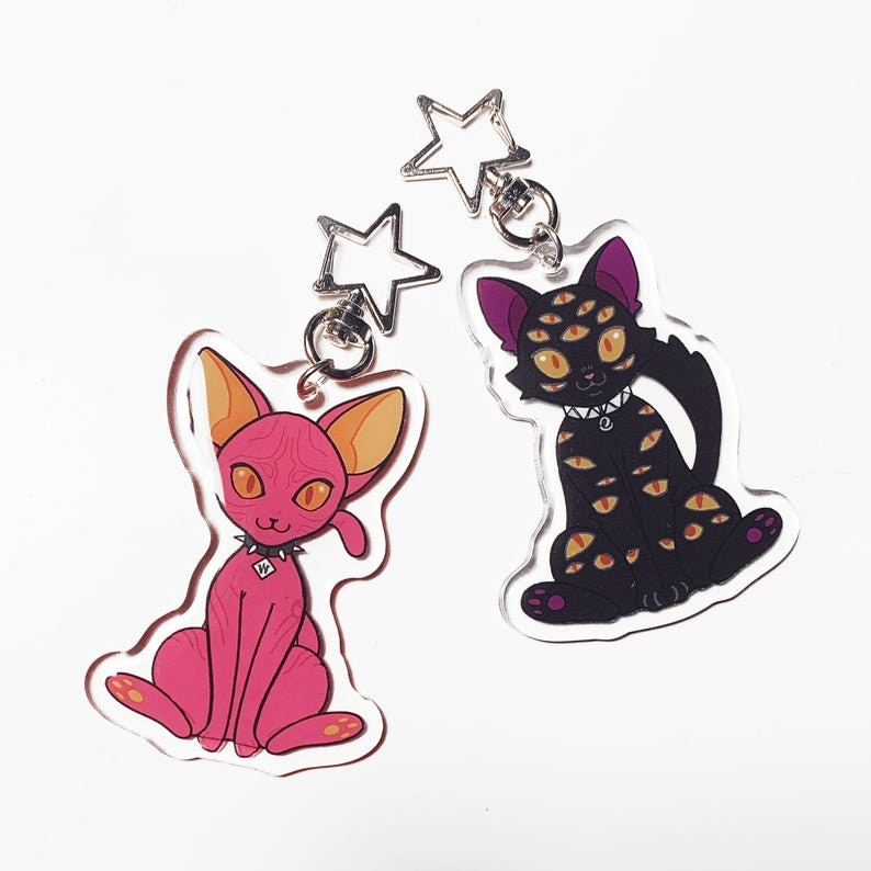 May include: Two acrylic keychains featuring cartoon cats. One cat is pink with orange eyes and the other is black with yellow eyes. Both cats have a black choker with a silver stud.