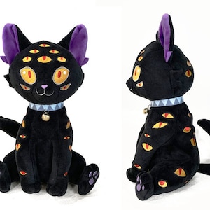 May include: Black plush cat with purple ears and a bell around its neck. The cat has multiple yellow eyes with red pupils.