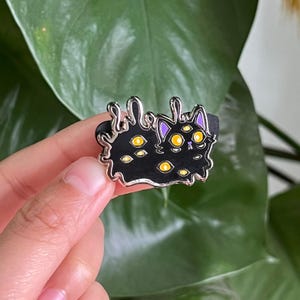 May include: Black enamel pin of a cat with multiple yellow eyes, melting into a puddle of black goo. The cat has purple ears and a white nose.
