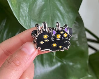 Puede incluir: Pin de esmalte negro de un gato con múltiples ojos amarillos, derritiéndose en un charco de baba negra. El gato tiene orejas moradas y una nariz blanca.