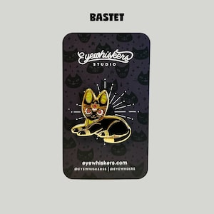May include: Gold-colored enamel pin featuring Bastet, the Egyptian goddess of cats, depicted as a black cat with a golden crown and red eyes. The pin is on a black background with a repeating pattern of stylized cats and stars. The text "Eye Whiskers Studio" and "eyewhiskers.com" are also visible.