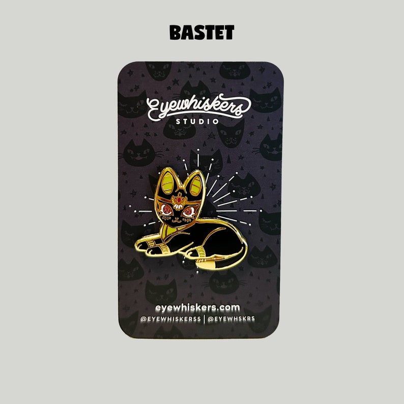 May include: Gold-colored enamel pin featuring Bastet, the Egyptian goddess of cats, depicted as a black cat with a golden crown and red eyes. The pin is on a black background with a repeating pattern of stylized cats and stars. The text "Eye Whiskers Studio" and "eyewhiskers.com" are also visible.