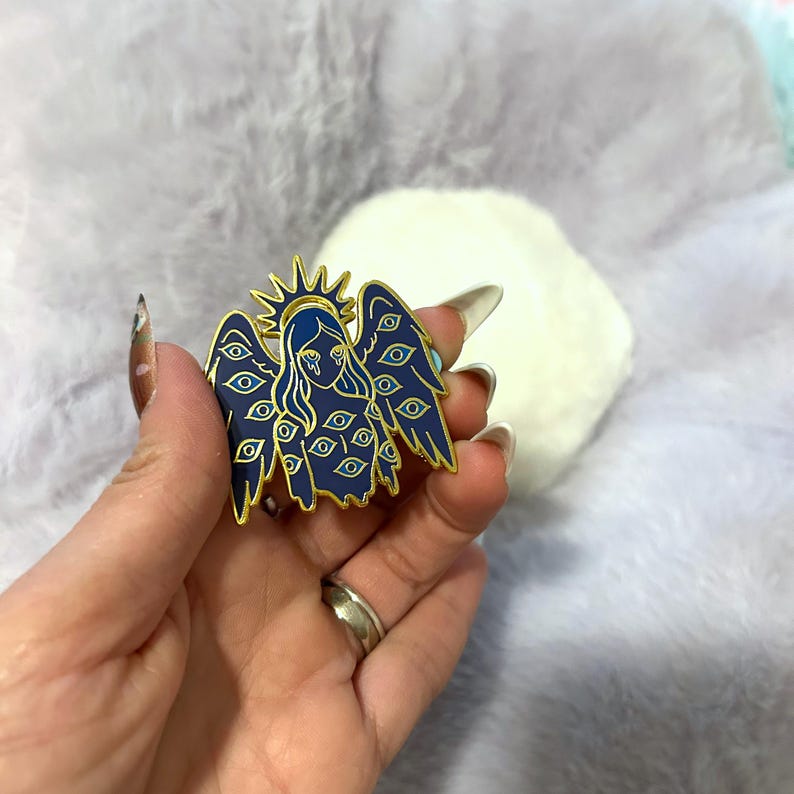 May include: A blue and gold enamel pin featuring an angel design with multiple eyes. The angel has a halo and is crying. The pin is held in a hand against a soft, light purple background.