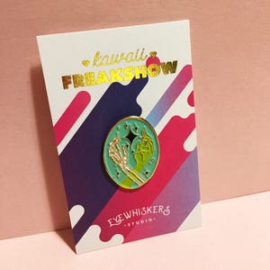 May include: A teal enamel pin with a gold outline featuring a skeleton hand holding a green hand with a star. The pin is on a white card with the text "kawaii FREAKSHOW" in gold lettering and "EYEWHISKERS STUDIO." in black lettering.