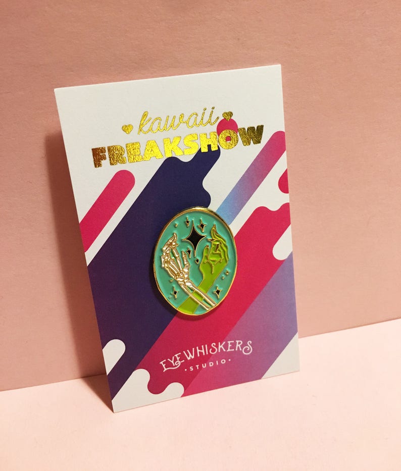 May include: A teal enamel pin with a gold outline featuring a skeleton hand holding a green hand with a star. The pin is on a white card with the text "kawaii FREAKSHOW" in gold lettering and "EYEWHISKERS STUDIO." in black lettering.