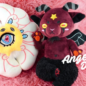 Puede incluir: Un conjunto de peluche de &aacute;ngel y demonio. El &aacute;ngel es una nube de peluche blanca con una cara amarilla y m&uacute;ltiples ojos. El demonio es un peluche burdeos con cuernos, alas y piernas negras. El texto "Be Not Afraid" y "Angel & Demon plush set" es visible en la imagen.