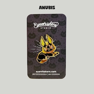 May include: Gold and black enamel pin featuring Anubis, the Egyptian god of the dead, with a cat-like face and a golden collar. The pin is on a black background with a white star pattern and the text "eyewhiskers studio" and "eyewhiskers.com".