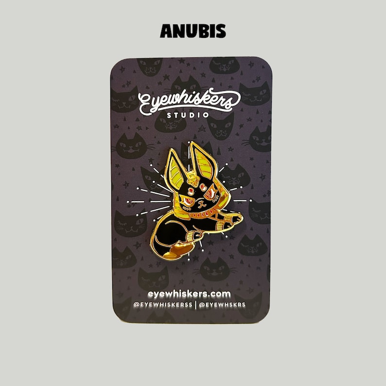 May include: Gold and black enamel pin featuring Anubis, the Egyptian god of the dead, with a cat-like face and a golden collar. The pin is on a black background with a white star pattern and the text "eyewhiskers studio" and "eyewhiskers.com".