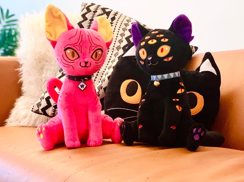 May include: Two plush cat toys, one pink with yellow ears and one black with purple ears and multiple eyes. Both cats are wearing collars with spikes and a charm. The toys are sitting on a brown couch.