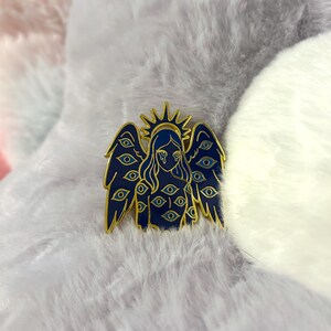 May include: A dark blue enamel pin featuring an angel with multiple eyes on the wings and body. The angel has a gold halo and gold outline. The eyes are blue with gold accents. The pin is set against a fluffy, light-colored background.