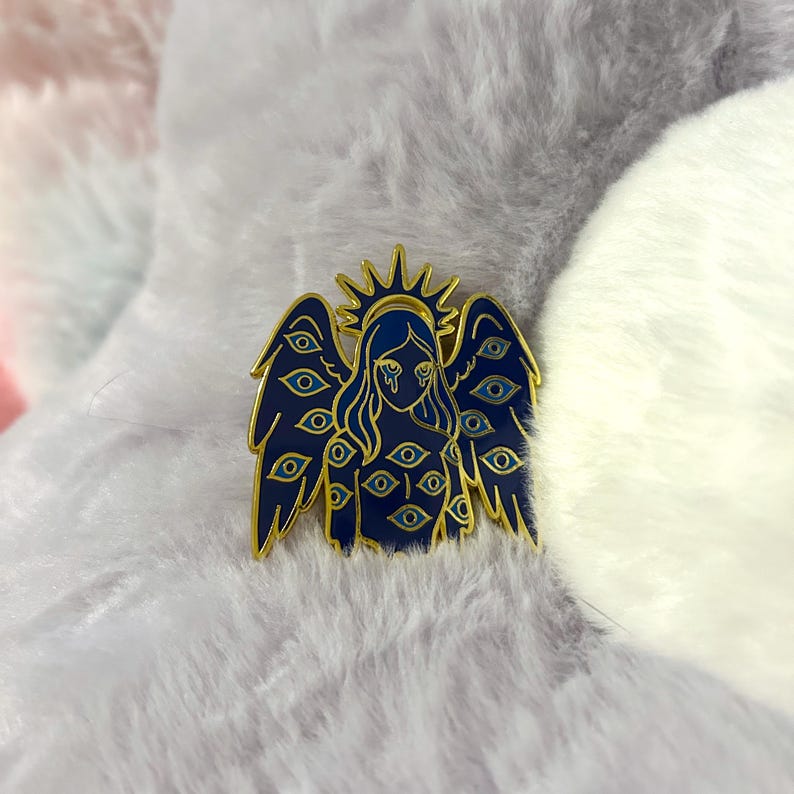 May include: A dark blue enamel pin featuring an angel with multiple eyes on the wings and body. The angel has a gold halo and gold outline. The eyes are blue with gold accents. The pin is set against a fluffy, light-colored background.