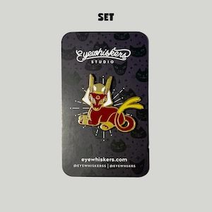 May include: Gold and red enamel pin featuring a stylized cat with a golden crown and a yellow tail. The pin is on a black background with a white cat pattern and the text "eyewhiskers studio" and "eyewhiskers.com".