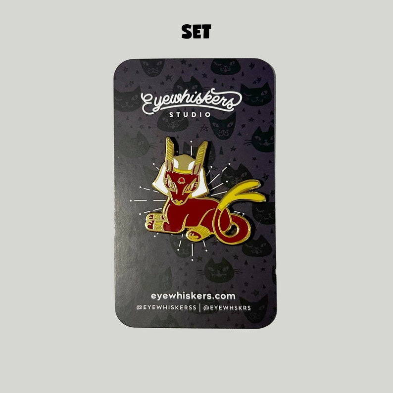 May include: Gold and red enamel pin featuring a stylized cat with a golden crown and a yellow tail. The pin is on a black background with a white cat pattern and the text "eyewhiskers studio" and "eyewhiskers.com".
