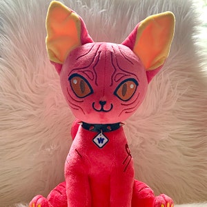 May include: Pink plush cat toy with a black studded collar and a white tag with the letter "W" on it. The cat has yellow ears and a detailed face.
