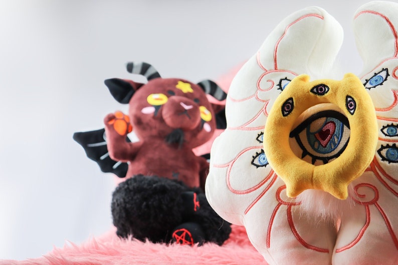 Puede incluir: Dos peluches, uno rojo con alas y cuernos negros, el otro blanco con ojos amarillos y azules y una boca grande.