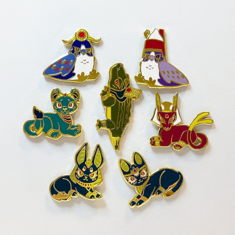 May include: Set of ten enamel pins featuring various Egyptian-themed designs. The pins are gold-toned and feature a variety of colors, including blue, red, green, and purple. The designs include a bird, a cat, a crocodile, a scarab beetle, and a pharaoh.