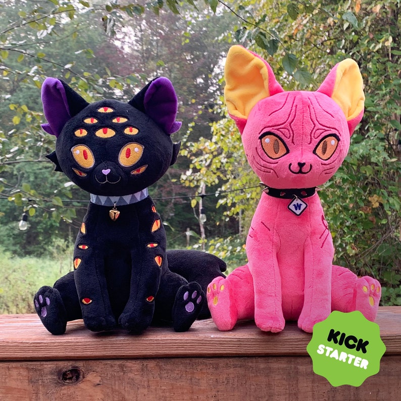 May include: Two plush cat toys. One is black with multiple eyes and a gold bell. The other is pink with yellow ears and a silver charm. Both have a black collar with a white letter.
