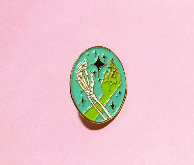 May include: A teal enamel pin with a skeleton hand and a green hand reaching towards a black star. The background is filled with small white stars.