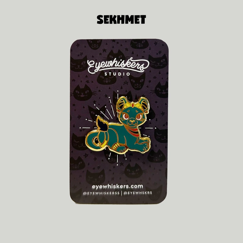 May include: Gold and teal enamel pin featuring a stylized cat with a crown, the text "SEKHMET" at the top, and "eyewhiskers STUDIO" and "eyewhiskers.com" at the bottom.