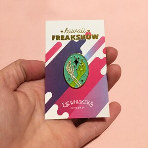 May include: A green enamel pin with a gold outline featuring a skeleton hand and a human hand holding a star. The pin is on a white card with the text "kawaii FREAKSHOW" and "EYEWHISKERS STUDIO."