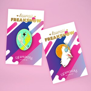 May include: Two enamel pins with a colorful background. The first pin features a green and yellow skeleton hand holding a star. The second pin features a white wolf howling at the moon. Both pins have the text "kawaii FREAKSHOW" and "EYE WHISKERS STUDIO."