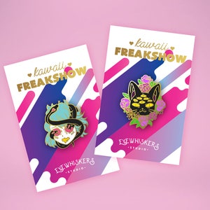 May include: Two enamel pins with a pink, purple, and blue background. The pins feature a black cat with multiple eyes and a woman with a snake wrapped around her head. The text on the cards reads "kawaii FREAKSHOW" and "EYEWHISKERS STUDIO."