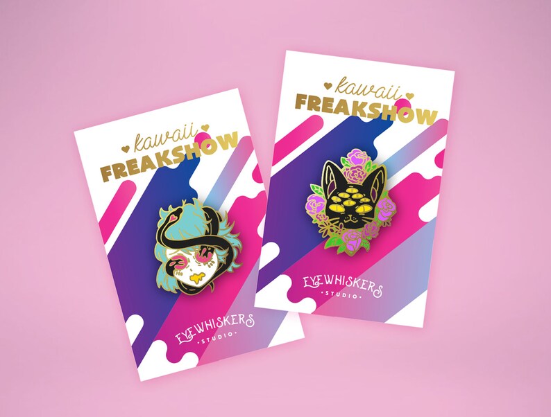 May include: Two enamel pins with a pink, purple, and blue background. The pins feature a black cat with multiple eyes and a woman with a snake wrapped around her head. The text on the cards reads "kawaii FREAKSHOW" and "EYEWHISKERS STUDIO."