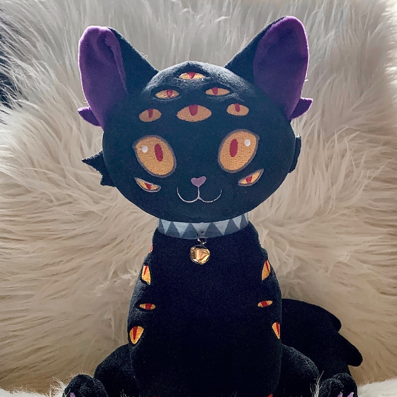 May include: Black plush cat toy with purple ears and five yellow eyes. The cat is wearing a black and white collar with a gold bell.