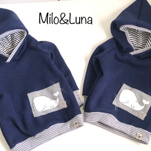 Könnte beinhalten: Zwei marineblaue Kapuzenpullover mit Wal-Grafik. Die Kapuzenpullover haben gestreiftes Futter in der Kapuze und an den Bündchen. Der Text "Milo & Luna" ist oben aufgedruckt.