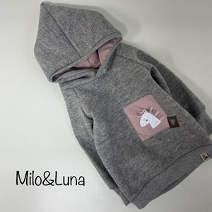 Könnte beinhalten: Grauer Kapuzenpullover mit rosa Innenfutter. Der Pullover hat einen Aufnäher mit einem weißen Einhorn-Design auf rosa Hintergrund. Der Markenname "Milo&Luna" ist unten links sichtbar.