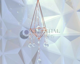 May include: A copper wire geometric crystal suncatcher with clear crystal drops hanging from it. The suncatcher is in the shape of a pyramid and is hanging from a white background.
