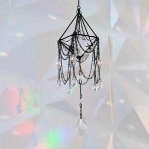 May include: A black metal chandelier with crystal drops and star-shaped accents hangs from a white ceiling. The chandelier casts rainbows on the white wall behind it.
